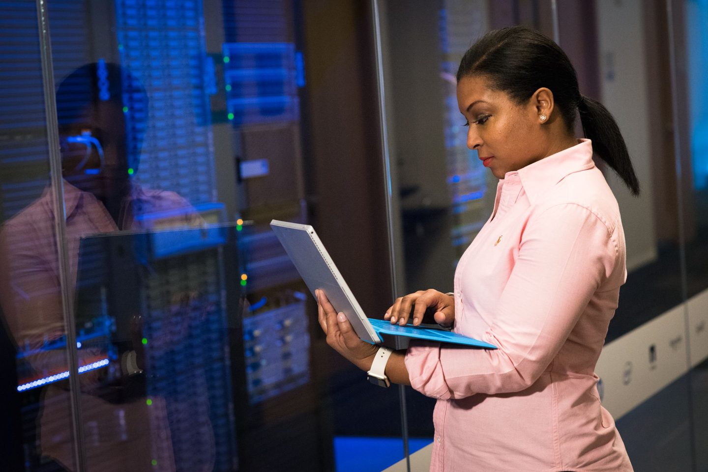 An engineer in a pink shirt typing on a laptop in a server room aisle